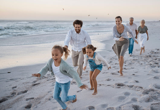 Familia multigeneracional corriendo y riendo en la playa, con los niños delante, seguidos por los padres y los abuelos.